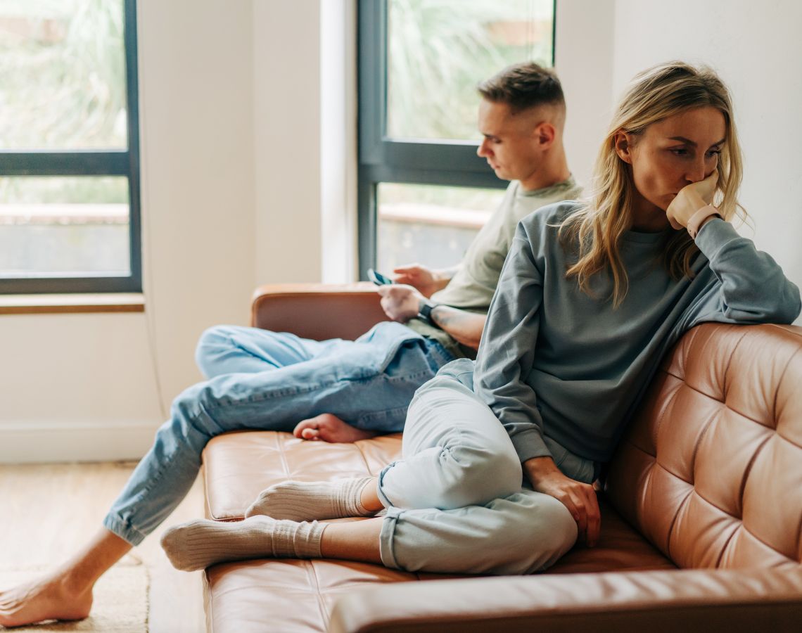 Paar in einer Beziehungskrise sitzt mit Abstand voneinander auf dem Sofa und stellt sich die Frage "Gehen oder bleiben"